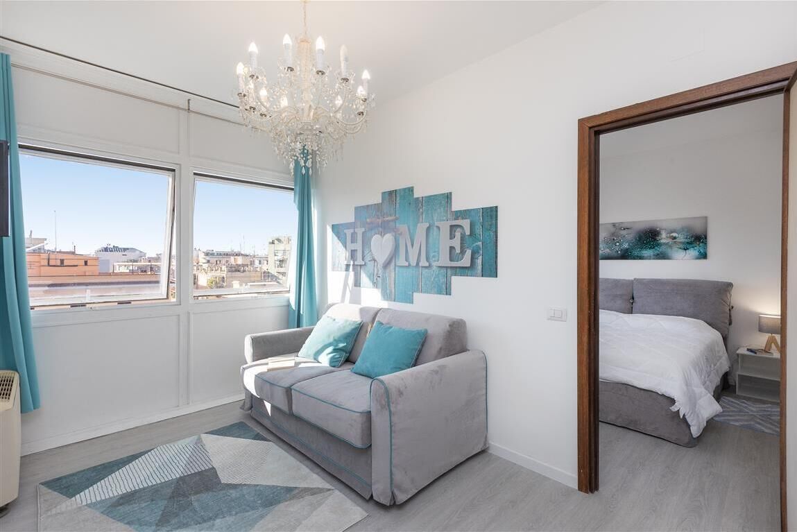 A modern bedroom with a grey sofa, a bed, a chandelier, and a large 'HOME' sign on the wall.