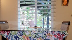 A dining table with a floral tablecloth, two chairs, a window with blinds, and a wall with a floral tapestry. - Large studio-prvt entry-prvt bathroom-option A/C (Papaikou)
