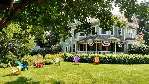 A large, two-story house with a porch decorated with American flags, surrounded by a well-maintained lawn and various Adirondack chairs.