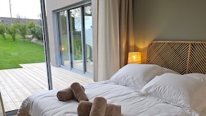 A bedroom with a large bed, a wooden headboard, and a view of a garden through a sliding glass door. - Villa with indoor pool 5 minutes walk from the beach (Plouguerneau)