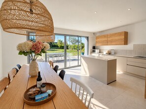 Et moderne køkken med en central ø, et spiseområde med et træbord og et stort vindue med udsigt over naturen.