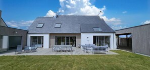 Una casa moderna con techo inclinado, grandes puertas de vidrio y una terraza con mobiliario exterior.