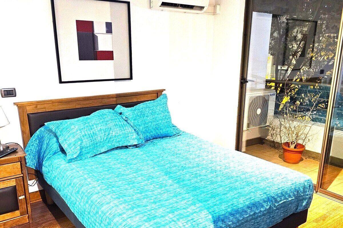 A bed with a blue bedspread and pillows, a wooden nightstand with a lamp, and a window with a view of trees and a balcony.