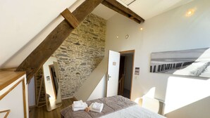 Ein Schlafzimmer mit Steinwand, Holzbalken, einem Bett mit Handtüchern und Meerblick durch ein Fenster.