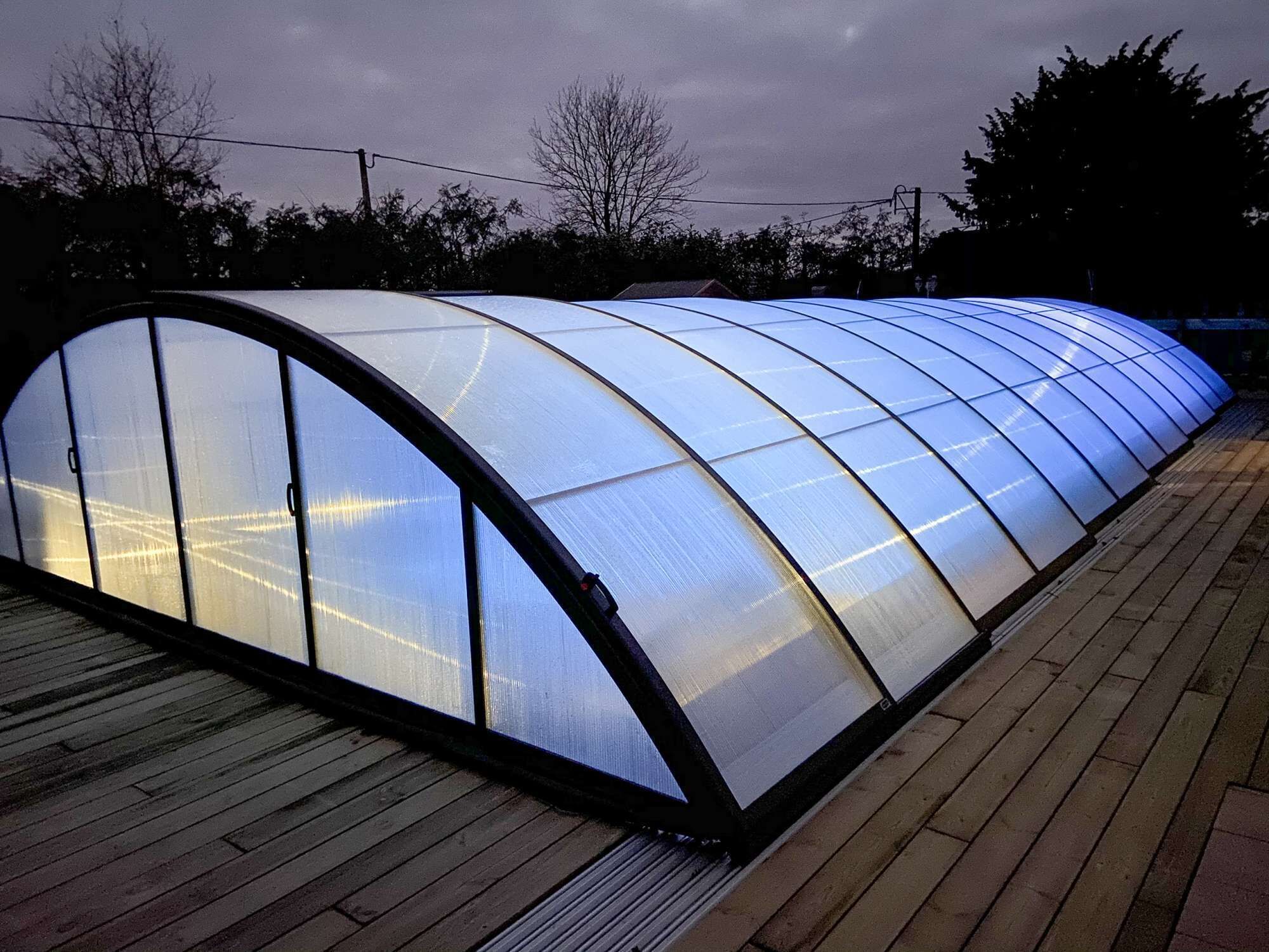 Una gran estructura de invernadero semitransparente con techo curvo, ubicada sobre una terraza de madera.