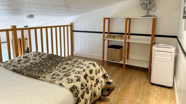 Un dormitorio con cama, estante de madera con libros, ventilador y calefactor.