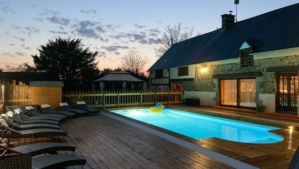 Ein Schwimmbecken mit Liegestühlen, einer Holzterrasse und einem Steinhaus mit dunklem Dach.