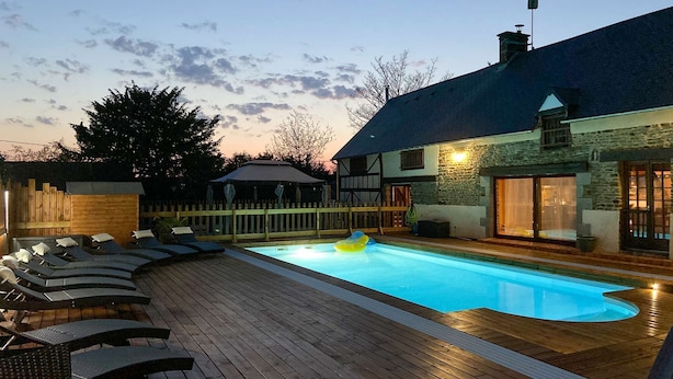 Ein Schwimmbecken mit Liegestühlen, einer Holzterrasse und einem Steinhaus mit dunklem Dach.