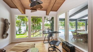 Una habitación amplia con ventanas grandes, techo de madera, zona de comedor con mobiliario de mimbre y vistas a la zona de la piscina exterior.