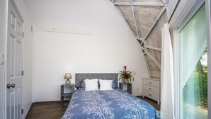 Un dormitorio con techo inclinado, una cama con colcha estampada de flores azules, una mesita de noche con lámpara, un aparador y una ventana con vistas al follaje.