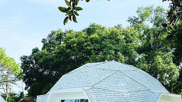 Un edificio moderno de forma abovedada y color blanco, con amplias ventanas, rodeado de abundante vegetación.