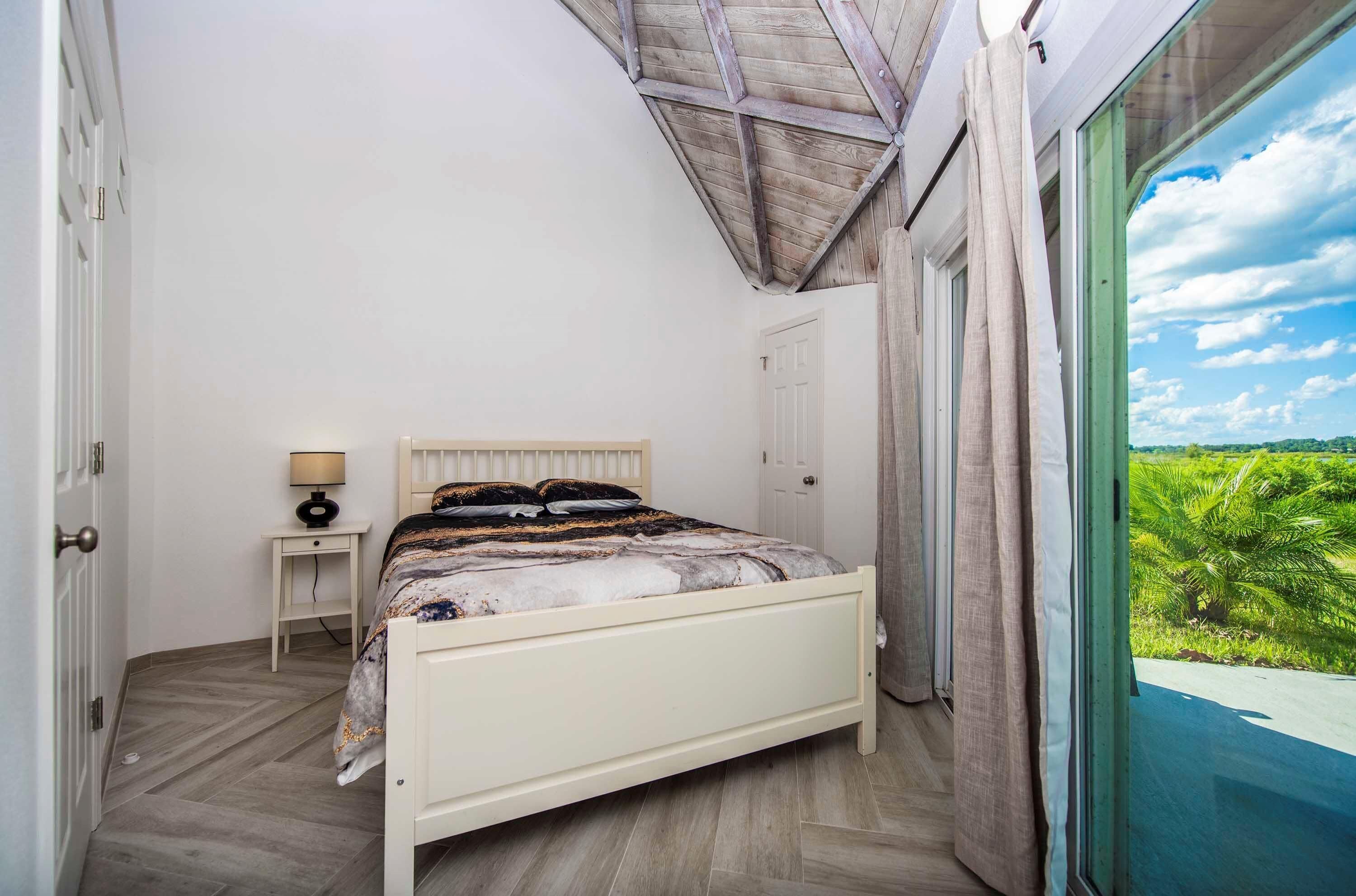 Un dormitorio con cama, mesita de noche, lámpara y vistas al verdor por una ventana.