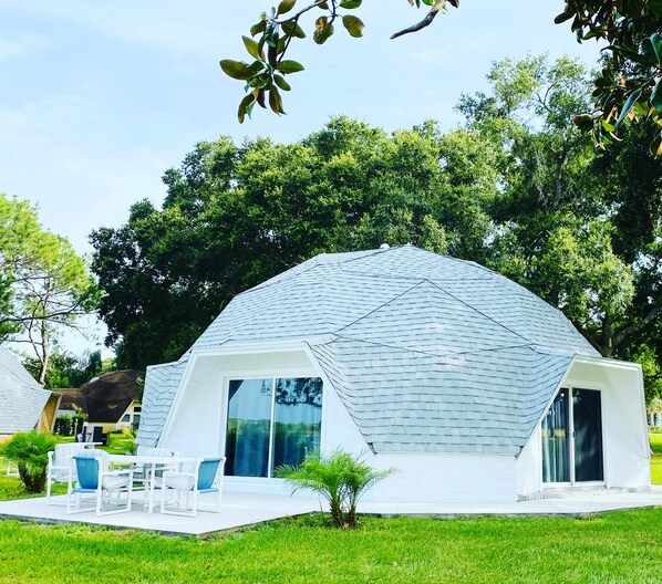 Una estructura de cúpula geodésica blanca con techo plano, rodeada de abundante follaje y árboles.