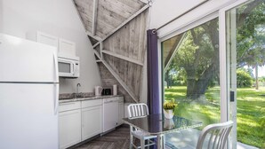 A modern kitchen with white cabinets, a microwave, and a coffee maker. There is a glass table with two chairs, and a view of a green garden through a large window. - Studio 01 - Lake Sumner (Clermont)