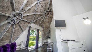 A room with a sloped ceiling, a flat-screen TV mounted on the wall, a desk with a chair, and a window with a view of greenery outside. - Studio 02 - Lake Sumner (Clermont)