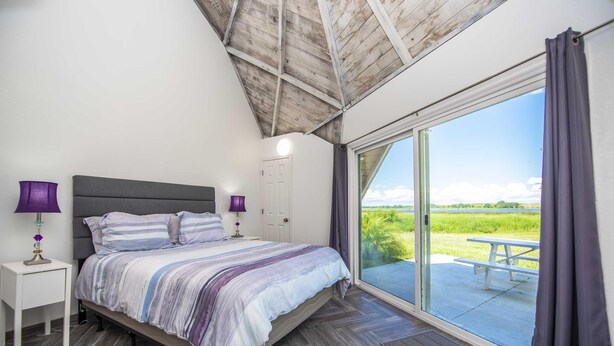 A bedroom with a large bed, two bedside tables with lamps, a white door, and a sliding glass door leading to a patio with a picnic table. - Studio 02 - Lake Sumner (Clermont)