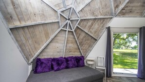 A modern bedroom with a sloped ceiling, a grey sofa bed, purple fluffy pillows, a white chair, and a view of a green lawn through a large window. - Studio 02 - Lake Sumner (Clermont)