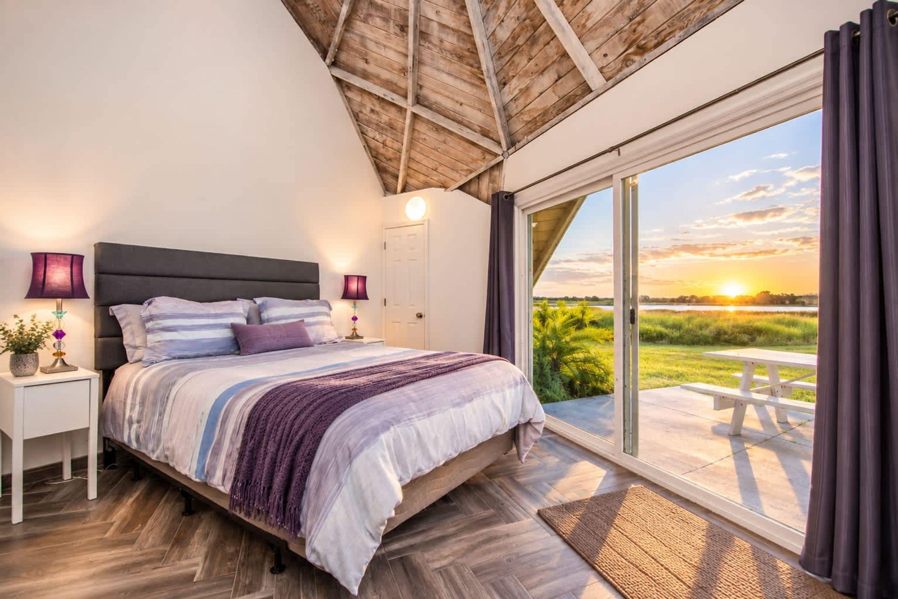 Quarto com cama grande, mesas de cabeceira, abajur e vista para o pôr do sol através de uma porta de correr.