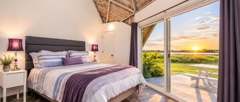 Un dormitorio con una cama grande, mesitas de noche, una lámpara y vistas al atardecer a través de una puerta corrediza de vidrio.