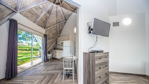 A room with a herringbone floor, a wooden ceiling, a dining table with chairs, a TV mounted on the wall, and a kitchen area visible through a glass door. - Studio 05 - Lake Sumner (Clermont)