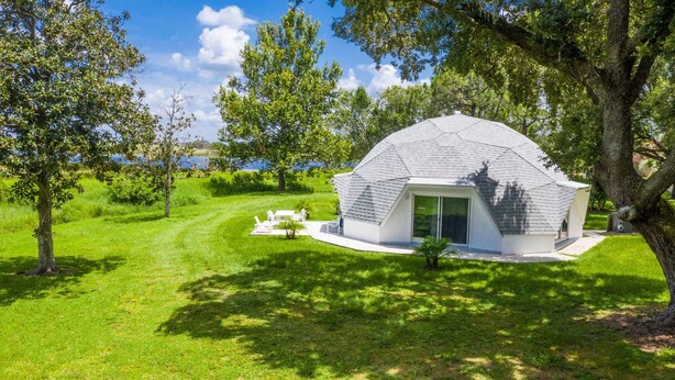 A modern, dome-shaped building with a white exterior, surrounded by lush greenery and trees, near a body of water. - Studio 05 - Lake Sumner (Clermont)