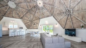 Ein geräumiges Loft mit hoher Holzbalkendecke, einem grauen Sofa, einem Fernseher und einer Küchenzeile mit Bar.