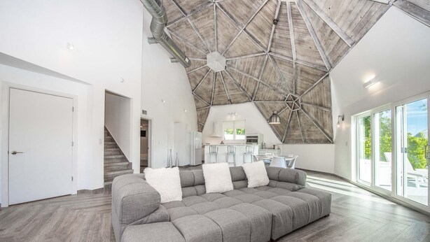 Un vaste espace de vie doté d’un grand canapé d’angle gris, d’un plafond en bois et offrant une vue sur l’extérieur grâce à de grandes fenêtres.