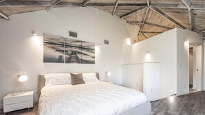 Une chambre avec un grand lit, des murs blancs et un plafond en bois. Il y a un tableau accroché au mur et une table de chevet avec une lampe.