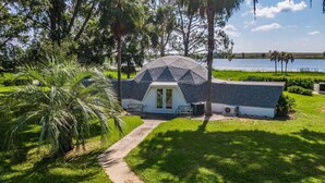 A small, single-story building with a sloped roof, surrounded by lush greenery and a body of water in the background. - Sumner Lake Tiny House - Lake Sumner (Clermont)
