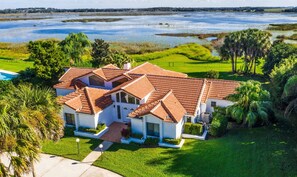 A large house with a red-tiled roof, surrounded by lush greenery and a body of water.