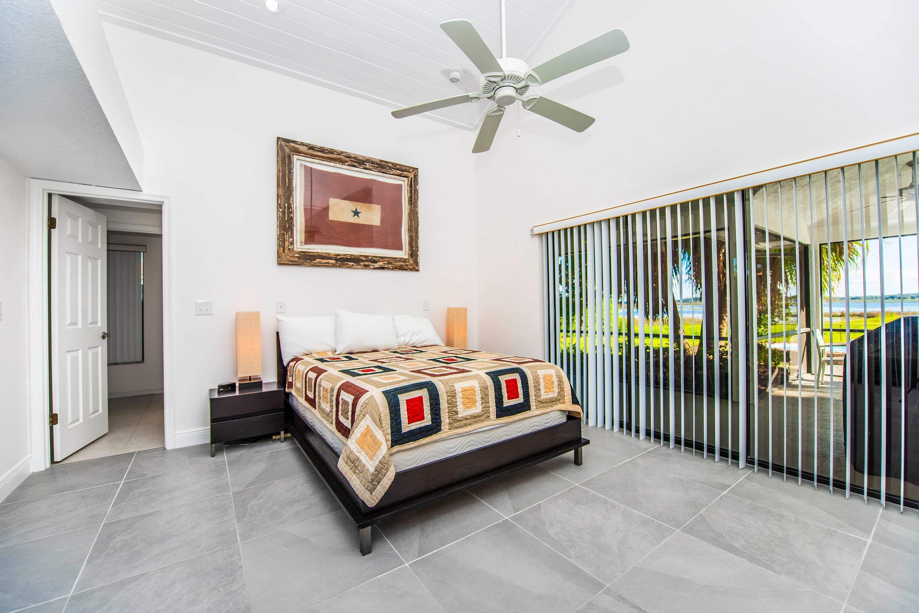 A bedroom with a bed, a ceiling fan, a framed picture, and a view through a window.