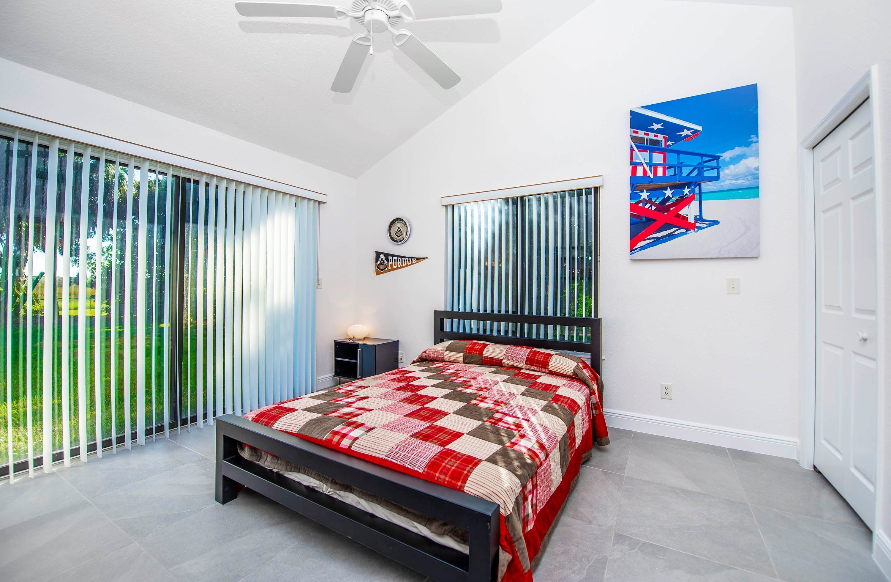 Ein Schlafzimmer mit einem Bett, einem Deckenventilator, einem Fenster mit Jalousien, einer Wanduhr und einem Gemälde.