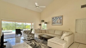 A spacious living room with a large sectional sofa, a coffee table, and a patterned rug. There is a ceiling fan, a painting on the wall, and a door to the right. - Swiss 101 - Lake Olsen (Clermont)