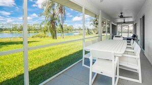 Um alpendre coberto com mesa e cadeiras, grandes janelas com vista para um lago e palmeiras.