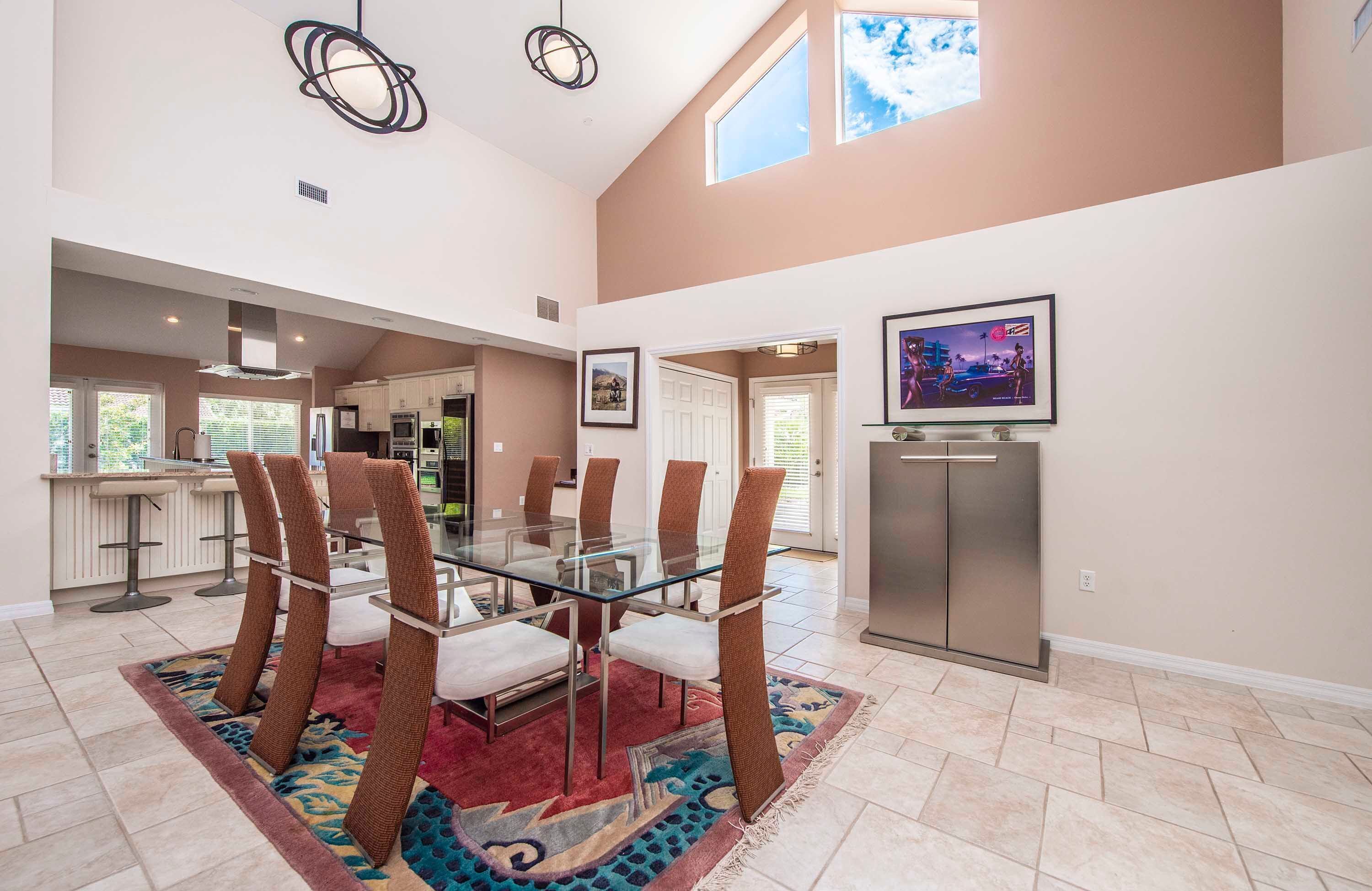 A spacious dining area with a glass table and brown chairs, a kitchen in the background, and a large window providing natural light.