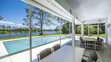 A poolside area with a table and chairs, large windows offering a view of a lake and trees.
