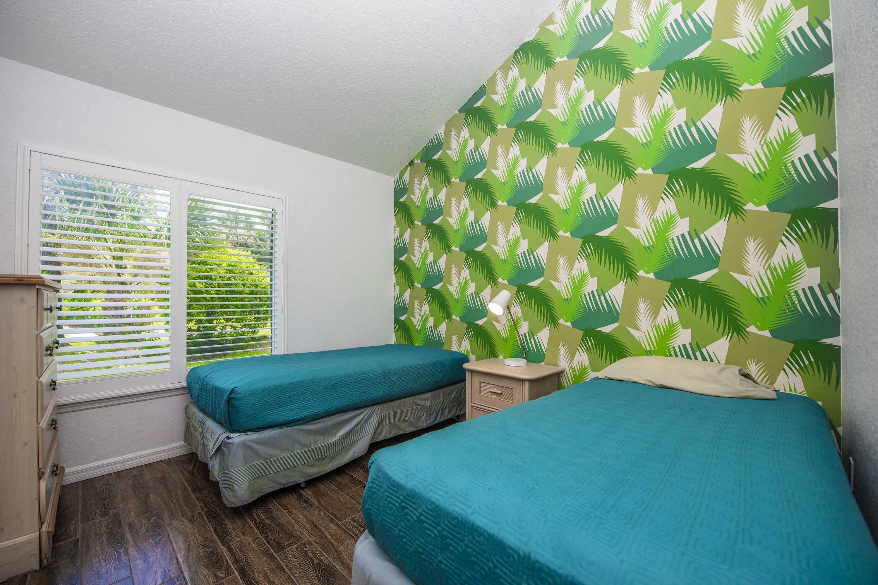 A room with two single beds, a window with blinds, a nightstand, and a wall with a leaf pattern.