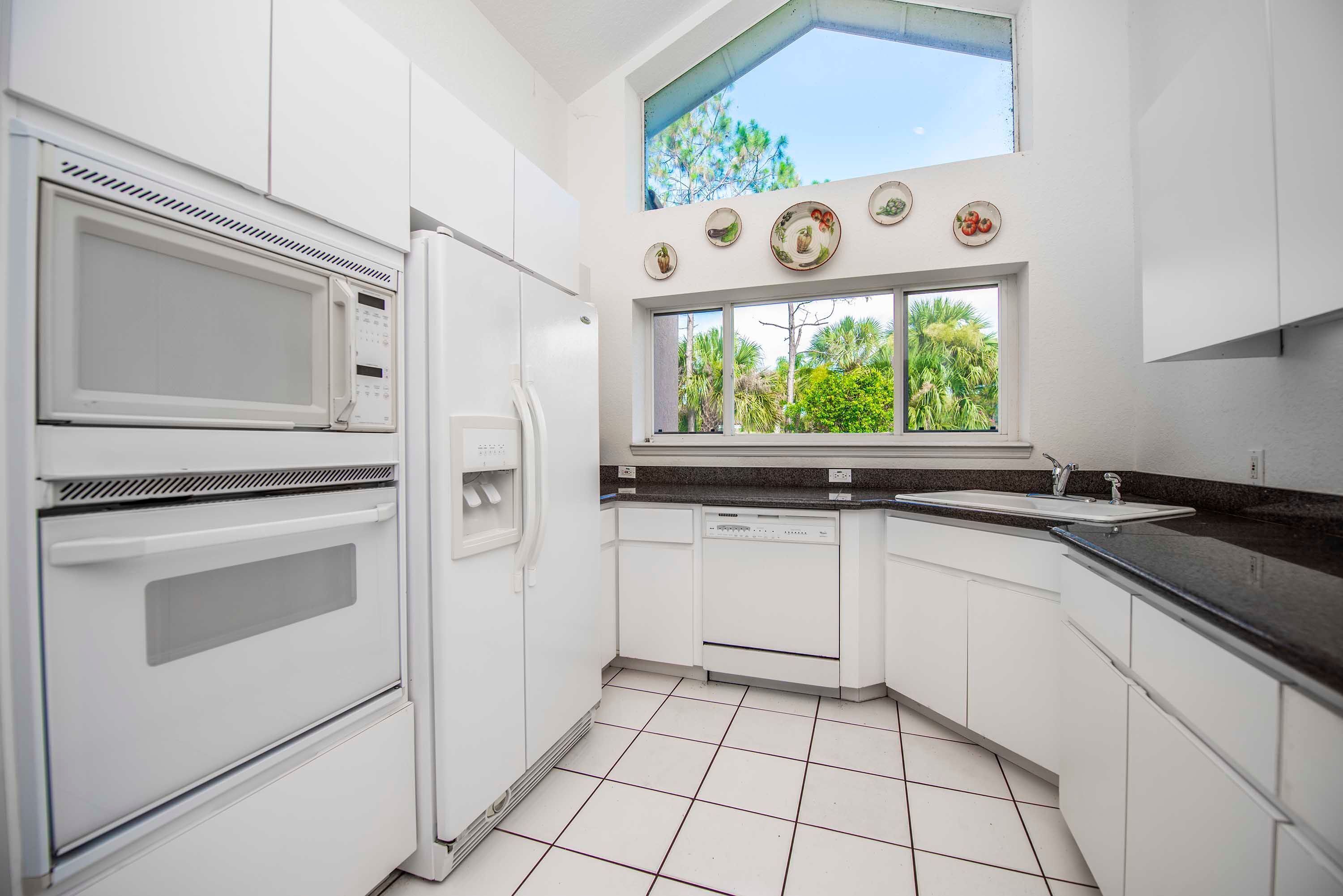 A modern kitchen with white cabinetry, a built-in oven, a microwave, a dishwasher, and a window with a view of greenery.