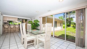 A dining area with a glass table and chairs, a kitchen in the background, and a view of a garden through large sliding doors. - Swiss 41 - Golf Course (Clermont)