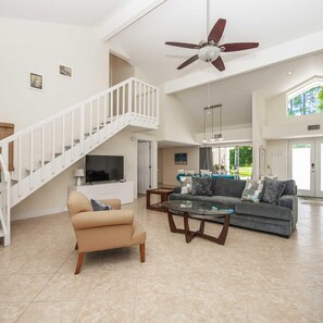A spacious living area with a high ceiling, a ceiling fan, a staircase, a flat-screen TV, a sofa, a coffee table, and a dining area with a view of the outdoors.