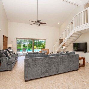 A spacious living room with a grey sofa set, a ceiling fan, a TV, and a staircase leading to an upper level.