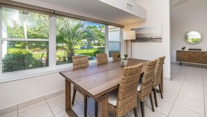 Uma sala de jantar com uma mesa grande e cadeiras de vime, janelas amplas com vista para o verde, uma cozinha moderna com armário de madeira e um espelho fixado na parede.