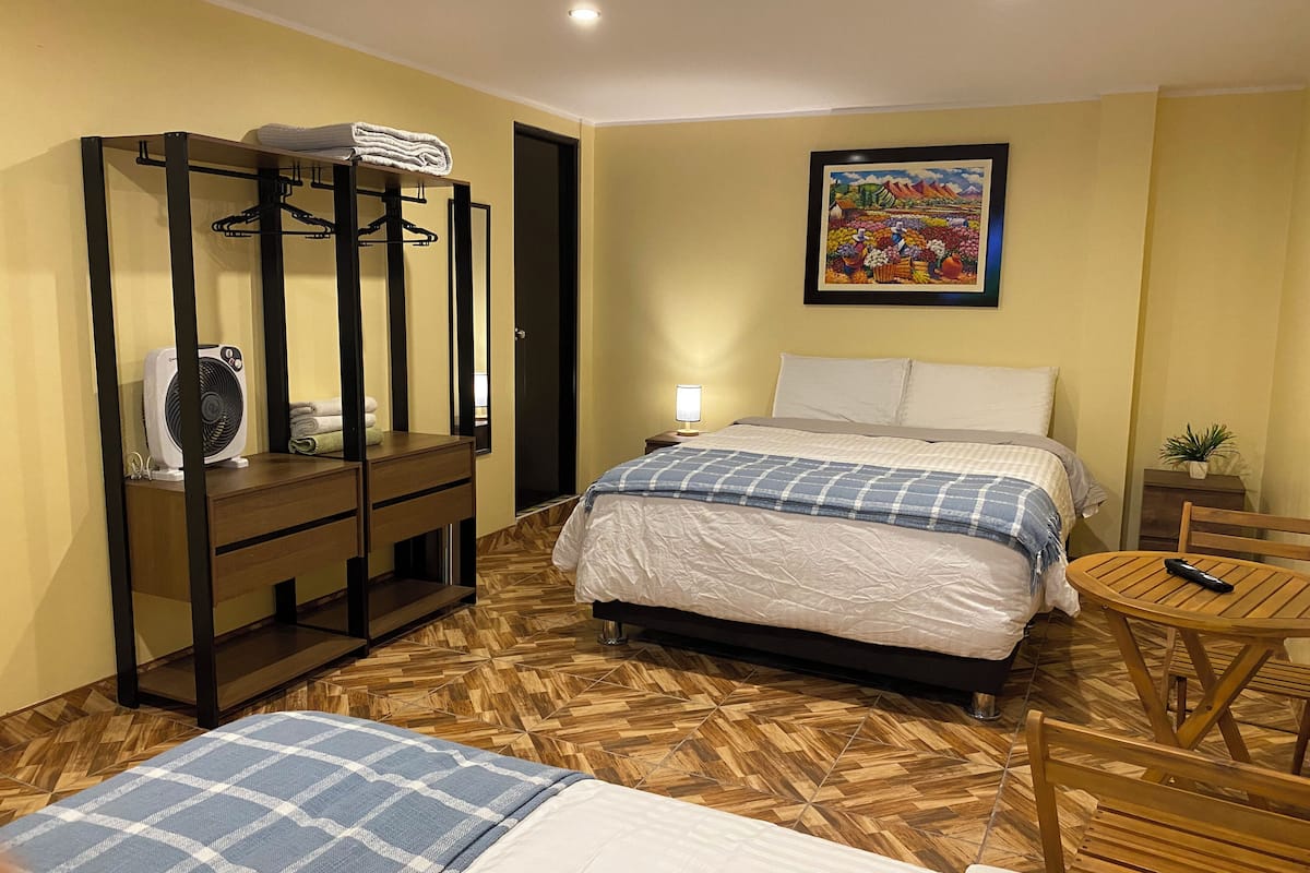 A hotel room with a bed, a desk with a fan, a chair, a small table, and a painting on the wall.