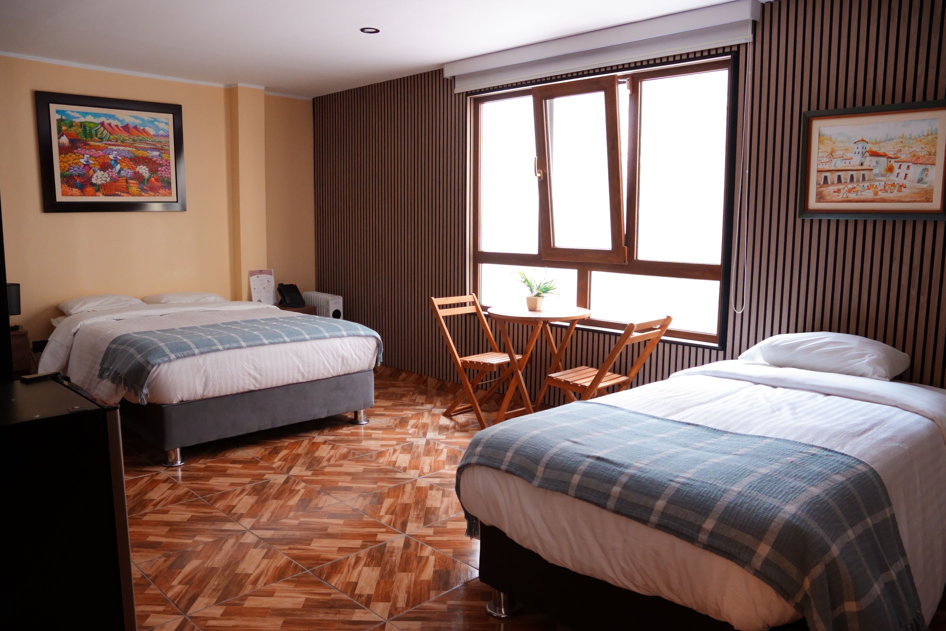 Quarto de hotel com duas camas, uma mesinha com cadeiras, uma planta em vaso e uma janela com persianas.