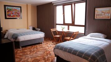 Quarto de hotel com duas camas, uma mesinha com cadeiras, uma planta em vaso e uma janela com persianas.