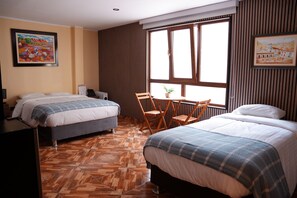 A hotel room with two beds, a small table with chairs, a potted plant, and a window with blinds. - Posada Miraflores (Lima)