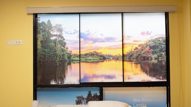 A hotel room with a bed, a nightstand with a lamp, and a large window with a scenic view of a lake and forest.