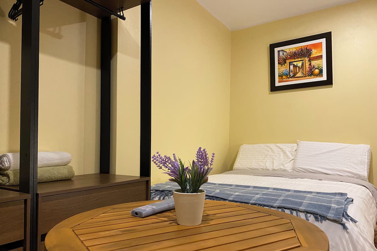 A bedroom with a bed, a bedside table with a remote control, a potted plant, and a painting on the wall.