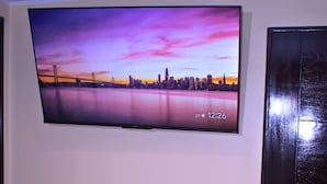 A room with a flat-screen TV mounted on the wall displaying a cityscape at sunset. There is a wooden table with two chairs and a small potted plant on it.