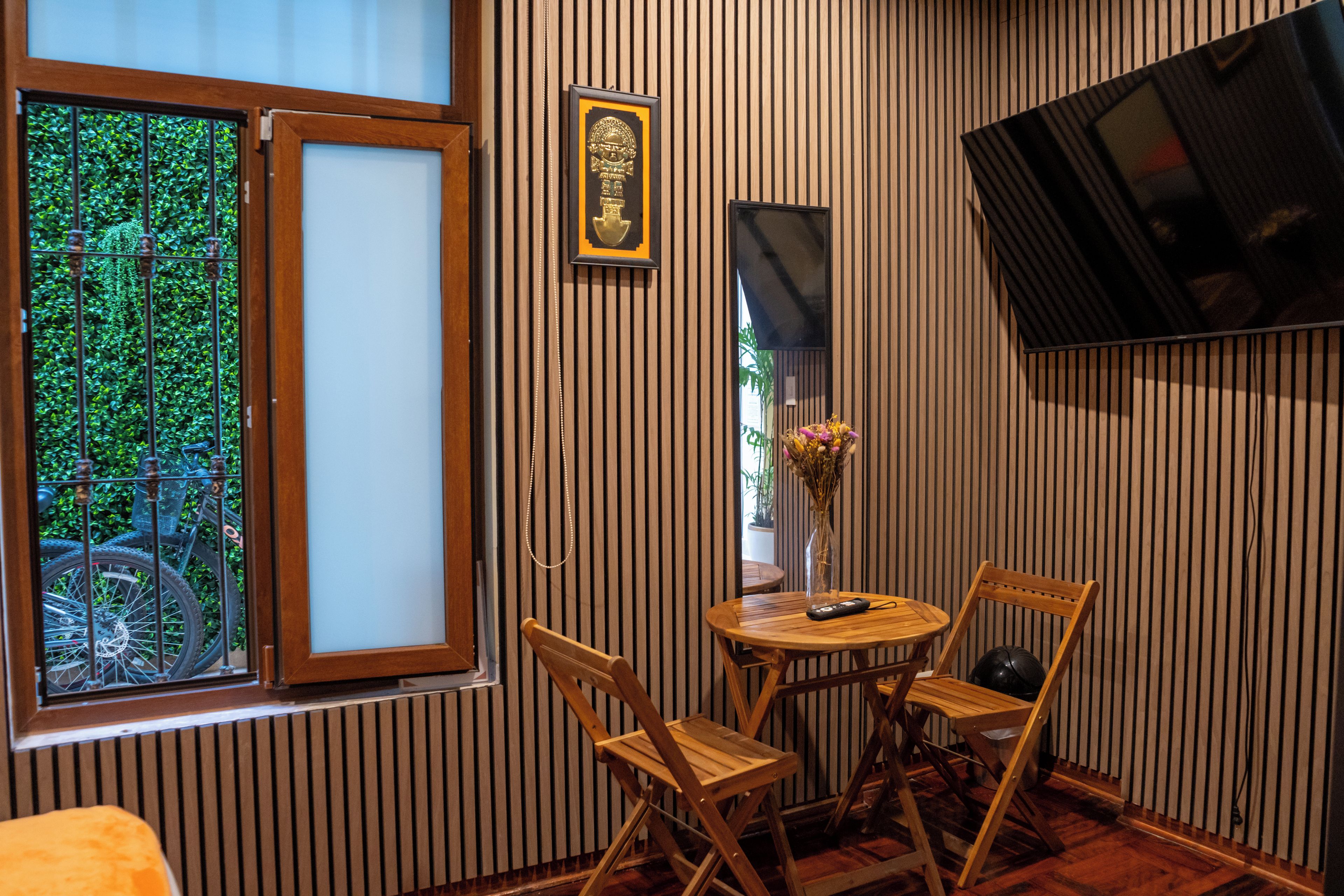 Una habitación con piso de madera, ventana con vista a follaje, una pequeña mesa con sillas y un televisor montado en la pared.